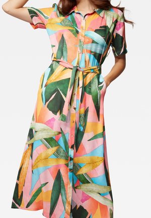 Femme portant une robe colorée à boutons, manches courtes, ceinture à la taille, avec un motif de feuilles tropicales en nuances de vert, rose, jaune et bleu.