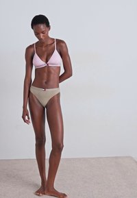 Bralette triangle rose clair avec bande logo, associé à un slip de bikini taille basse beige. Tissu doux, couverture minimale, design simple.