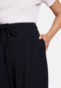 Pantalon taille haute bleu marine avec ceinture à nouer, doté de poches latérales et d'une texture de tissu lisse et fluide.