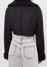 Svart kort jacka med en texturerad, plyschig krage och ärmslut. Slät material med en strukturerad design, ihopparad med högmidjade ljusa jeans.