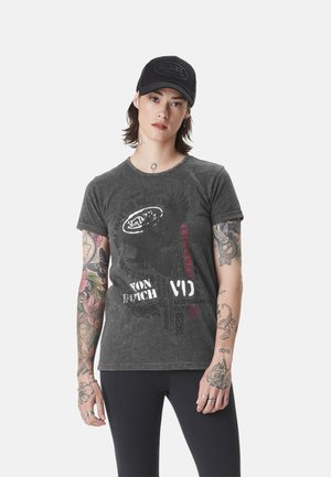 Personne avec des tatouages sur les deux bras portant une casquette noire Von Dutch et un T-shirt graphique gris foncé avec un legging noir sur un fond uni.