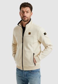 Veste zippée beige avec un motif matelassé texturé sur le devant supérieur, fermeture éclair noire, poches latérales et accents de marque sur la poitrine et la manche.