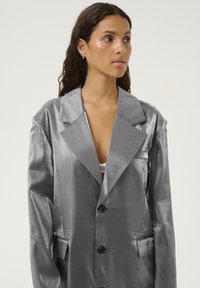 Silver satin blazer with a structured design, notched lapels, and six buttons. Features two front pockets and a slightly glossy texture.
