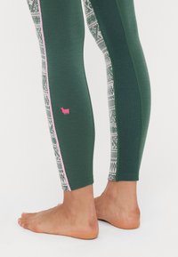Green leggings with textured fabric, featuring a patterned side panel and pink accent stripe, complemented by a small pink logo detail.