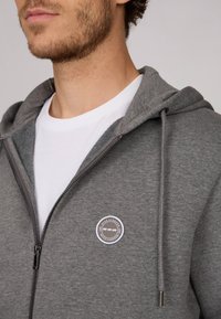 Un sweat à capuche gris zippé avec un cordon de serrage, présentant un patch logo circulaire sur la poitrine gauche et une texture lisse et douce.