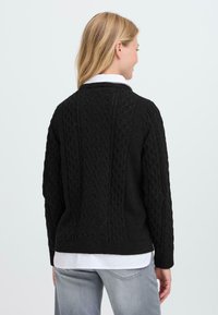 Maglione nero lavorato a maglia con un motivo a treccia testurizzato, scollo rotondo, maniche lunghe e vestibilità rilassata, abbinato a una camicia bianca sotto.