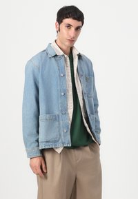 Light blue denim jacket featuring a button front, two front pockets, and a green logo. Worn over a plaid shirt and green top. Tan trousers.
