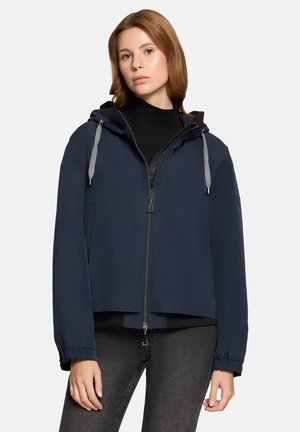 Woman wearing navy blue hooded zip-up jacket with black shirt and dark jeans, standing against a plain light background.