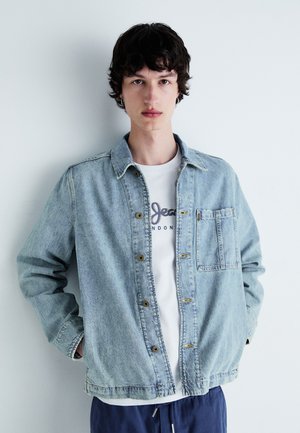 Light blue denim jacket with a collared neckline and front pocket, layered over a white t-shirt featuring blue text, simple design.