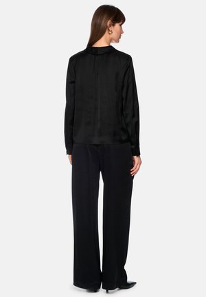 Woman with long brown hair wearing a long-sleeve black blouse and loose black pants, standing facing away on a white background.