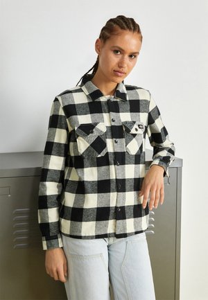 Camicia di flanella a scacchi in bianco e nero con colletto tradizionale, chiusure a bottone, due tasche frontali, indossata sopra jeans azzurri chiari.