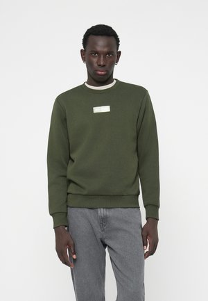 Homme portant un sweat-shirt vert olive avec un petit logo rectangulaire et un jean gris clair, debout contre un fond blanc uni.