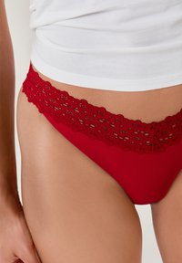 String rouge avec une taille en dentelle festonnée, ornée de motifs floraux complexes. Le tissu lisse se fond avec le teint en dessous de la taille.