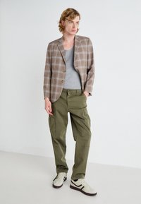 Brown plaid blazer with two buttons, gray crew neck shirt, olive cargo pants, and beige sneakers with dark accents. Textured fabric details visible.