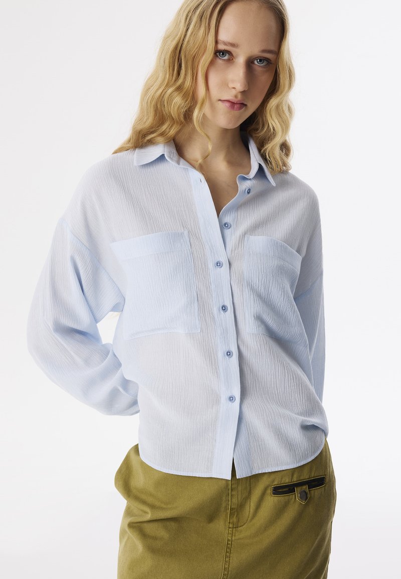 Camisa de botón azul claro con una tela texturizada, dos bolsillos frontales y un corte relajado, combinada con una falda verde oliva con un acento negro.
