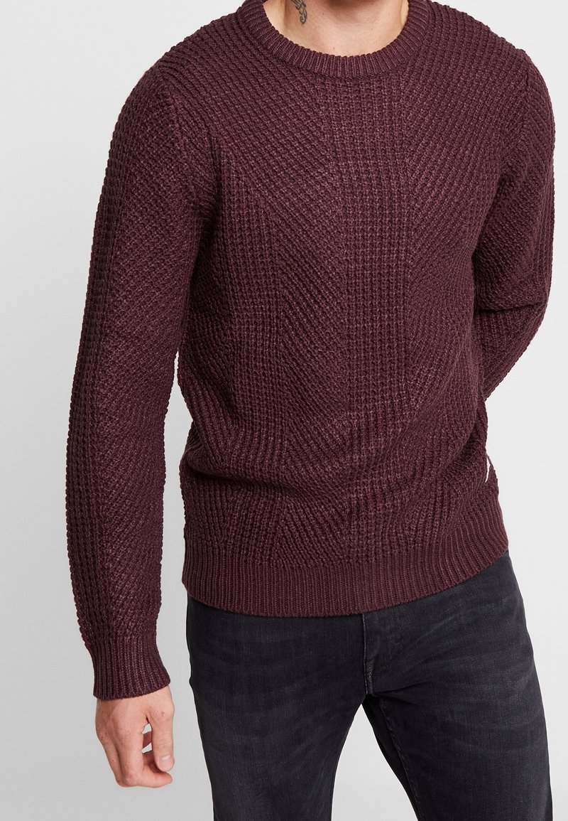 Uomo che indossa un maglione lavorato a maglia di un viola scuro e jeans neri, in piedi con una mano rilassata al suo fianco.