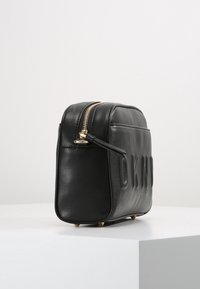 Borsa cosmetica rettangolare in pelle nera con zip dorata e scritta in rilievo sul lato, posizionata su una superficie bianca.