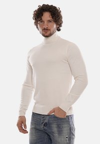 Maglione bianco a collo alto con maniche lunghe, design aderente, texture liscia, indossato con jeans chiari strappati.