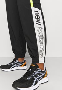 Osoba nosící černé tepláky New Balance s bíl�ými a žlutými pruhy, doplněné černými a oranžovými běžeckými botami Asics.