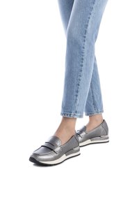 Mocasines metálicos grises con suela plana, que presentan una correa decorativa y un ligero tacón, combinados con jeans rectos de color azul claro.