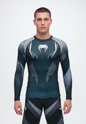 Un homme se tient face à l’avant, vêtu d’un haut de compression à manches longues bleu canard foncé et d’un legging de sport assorti noir et gris.
