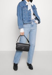 Sort håndtaske med sølvnitter, rektangulær form og et lille håndtag. Bæres med en lyseblå denimjakke og jeans. Sorte ankelstøvler fuldender looket.