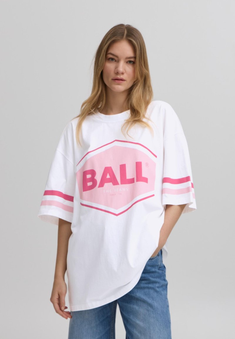 Camiseta blanca de gran tamaño con logo "BALL" en rosa, mangas cortas con rayas rosas y un ajuste relajado. Llevada con jeans azul claro.
