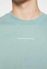 T-shirt in cotone verde chiaro con collo rotondo e maniche corte. Presenta la scritta ricamata bianca "SANDRO PARIS" sul petto.