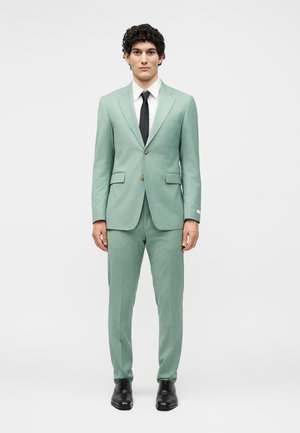 Άνδρας που στέκεται με το σώμα προς τα εμπρός, φοράει ένα κοστούμι σε μέντα πράσινο χρώμα, λευκό πουκάμισο, μαύρη γραβάτα και μαύρα δερμάτινα παπούτσια, πάνω σε ένα απλό λευκό φόντο.