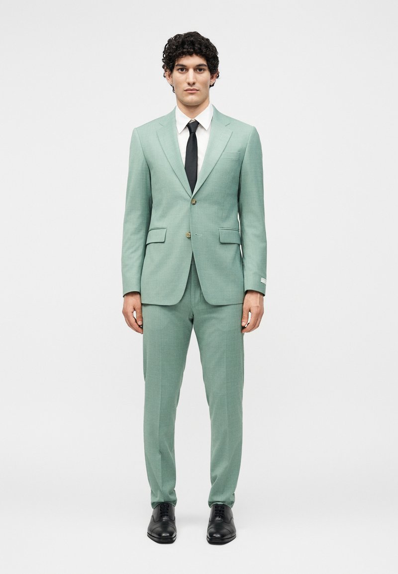 Άνδρας που στέκεται με το σώμα προς τα εμπρός, φοράει ένα κοστούμι σε μέντα πράσινο χρώμα, λευκό πουκάμισο, μαύρη γραβάτα και μαύρα δερμάτινα παπούτσια, πάνω σε ένα απλό λευκό φόντο.