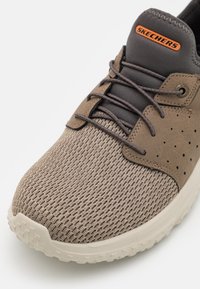 Zapatilla para hombre de Skechers en beige con parte superior de malla, detalles en gris oscuro, perforaciones y suela de goma texturizada.
