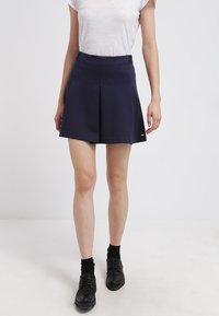 Gonna plissettata blu navy realizzata in tessuto liscio, con una fascia nera in vita e un logo in evidenza. Indossata con una maglietta bianca e scarpe nere.