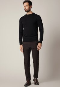 Mężczyzna stojący w czarnym swetrze, ciemnobrązowych prostych spodniach i ciemnobrązowych skórzanych półbutach na beżowym, jednolitym tle.