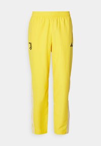Pantalones deportivos amarillos con cinturilla elástica, que presentan logotipos negros en el muslo izquierdo y franjas blancas a lo largo de las piernas.
