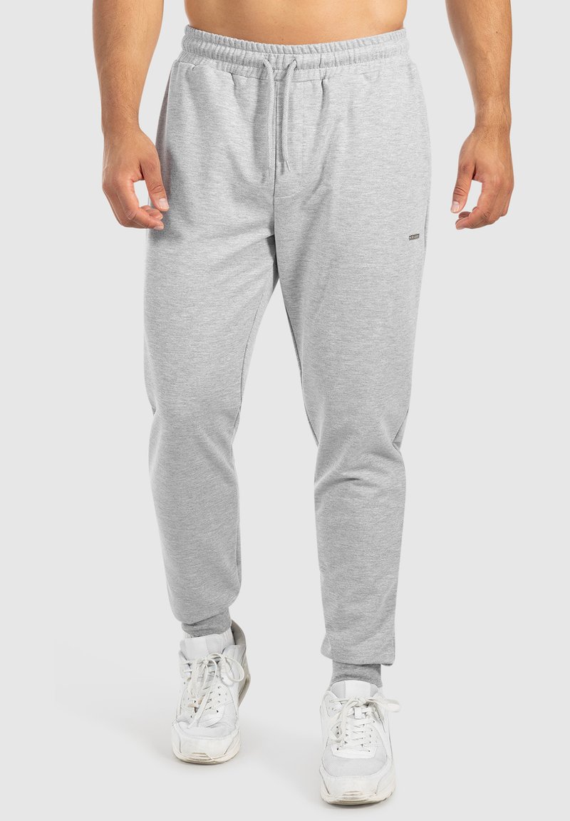 Pantalons de survêtement en coton gris avec taille à cordon, design à jambes fuselées et petit logo sur le côté. Portés avec des baskets blanches.