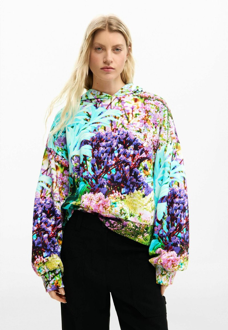 Blonde woman wearing a colorful floral print hoodie with purple, pink, and green hues, paired with black pants, standing against white background.