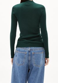 Maglia lunga a maniche lunghe verde scuro con colletto alto, abbinata a jeans blu a vita alta con un classico design a cinque tasche.