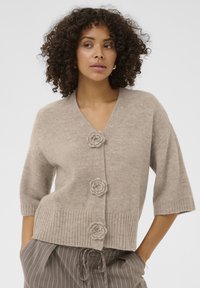 Beige kortærmet cardigan med v-hals, med tre blomsterformede knapper og ribstrikket bundkant. Blød tekstur og afslappet pasform.