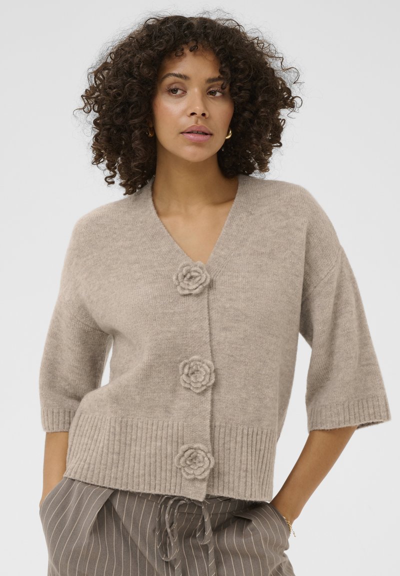 Beige kortærmet cardigan med v-hals, med tre blomsterformede knapper og ribstrikket bundkant. Blød tekstur og afslappet pasform.