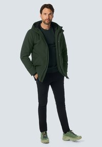 Donkergroene waterdichte jas met capuchon, gecombineerd met een zwarte trui en broek, aangevuld met lichtgroene sportschoenen.