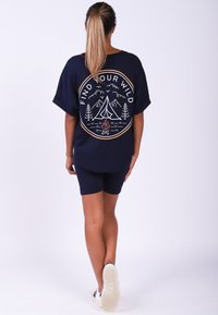 Marineblauw oversized t-shirt met een cirkelvormige campingafbeelding met bergen, bomen en een vuur. Gepaard met marineblauwe shorts en witte sneakers.