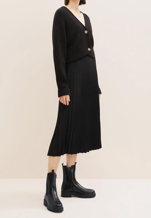 Frau trägt eine schwarze V-Ausschnitt-Strickjacke mit zwei Knöpfen, einen plissierten, knielangen schwarzen Rock und schwarze Stiefeletten mit groben Sohlen.