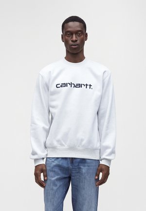 Άνδρας που φοράει λευκό φούτερ Carhartt και μπλε τζιν, στέκεται μπροστά σε απλό ανοιχτό γκρι φόντο.