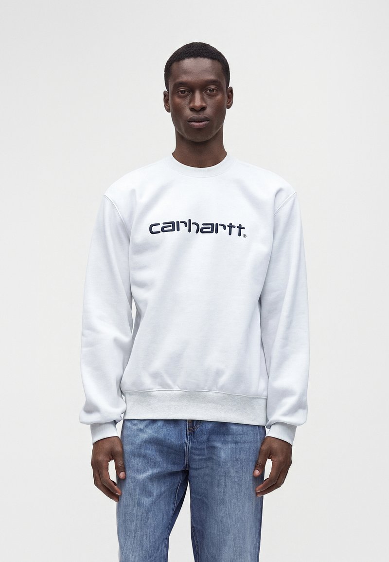 Άνδρας που φοράει λευκό φούτερ Carhartt και μπλε τζιν, στέκεται μπροστά σε απλό ανοιχτό γκρι φόντο.