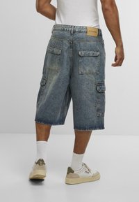 Denim cargo kratke hlače v izbledeli modri barvi z več žepi, ohlapnim fitom in šivanim robom. Imajo blagovno znamko iz rjave usnje na zadnji strani.