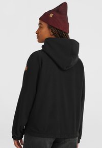 Black fleece jacket with a hood, featuring elastic cuffs and a leather logo patch on the left shoulder. Paired with a burgundy ribbed beanie.