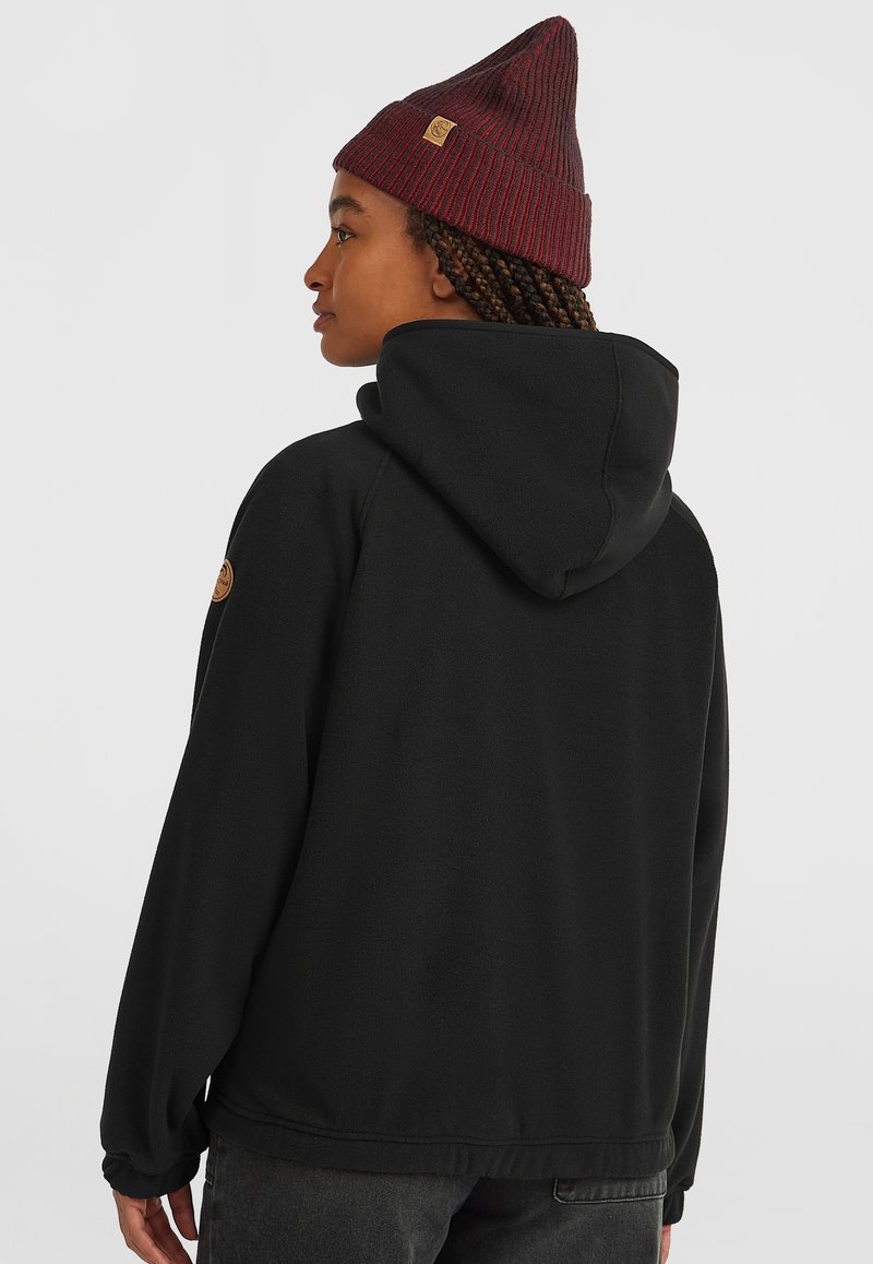 Schwarze Fleecejacke mit Kapuze, mit elastischen Bündchen und einem Lederlogo-Patch auf der linken Schulter. Kombiniert mit einer weinroten gerippten Beanie.