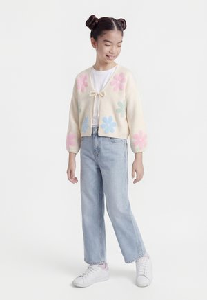 Ragazza giovane con doppio chignon, indossa un cardigan crema con fiori rosa e blu, jeans azzurri chiari e scarpe da ginnastica bianche.