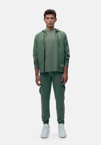 Grünes Sport-Outfit bestehend aus einer Zip-Hoodie und passenden Cargo-Hosen, mit einer weichen Textur und weißen Sneakers. Rote Kordeln betonen die Kapuze.