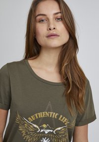 Oliven grøn kortærmet T-shirt med et slidt grafisk print af vinger og bjerge, med teksten "AUTHENTIC LIFE" i fed skrift.